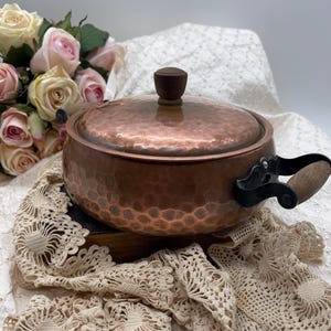 May include: A copper pot with a hammered texture, a wooden handle, and a matching lid with a wooden knob. The pot sits on a wooden base, with a cream-colored lace doily and a bouquet of pink and cream roses in the background.
