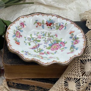 May include: A white, scalloped-edge dish with a floral and bird design. The dish has a gold-colored rim and is filled with pink, blue, and red flowers. The dish is placed on top of a stack of antique books.