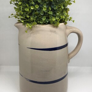 Puede incluir: Una jarra de cer&aacute;mica blanca con rayas azules, llena de una planta artificial verde.