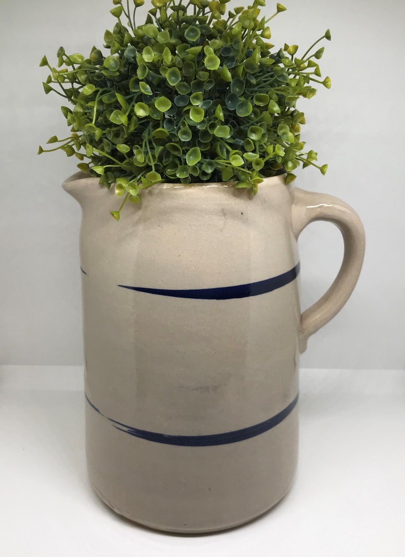 Puede incluir: Una jarra de cer&aacute;mica blanca con rayas azules, llena de una planta artificial verde.