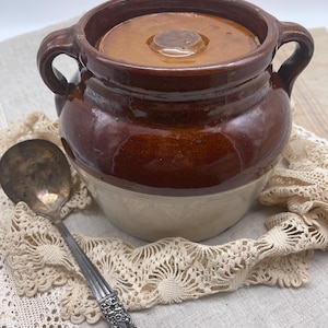 May include: A brown and white glazed ceramic crock with a lid and two handles. The crock is sitting on a white lace doily.