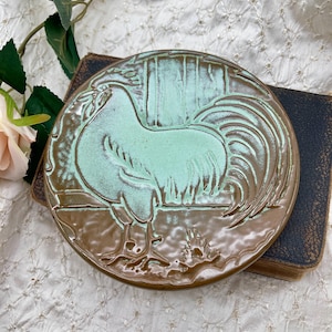 May include: A round ceramic tile featuring a raised relief design of a rooster. The rooster is finished in a light mint green glaze, while the background and border are a glossy brown. The surface has a textured, hand-crafted appearance.