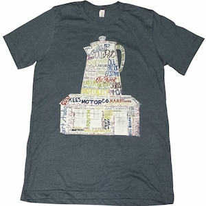 May include: A dark gray t-shirt featuring a graphic of a coffee pot and a building, both filled with various words. The design is in shades of white, yellow, and red. The shirt is made of a soft material.