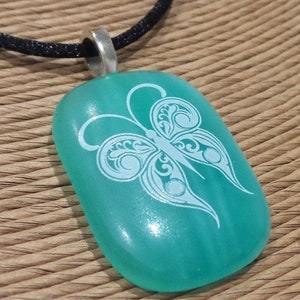 Puede incluir: Colgante turquesa con diseño de mariposa blanca. El colgante, de forma rectangular con bordes redondeados, cuelga de un cordón negro. La mariposa presenta intrincados diseños en las alas. El colgante es de material liso y brillante.