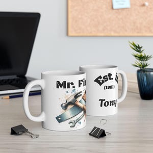May include: Two white ceramic mugs featuring a graphic of a hammer, saw, and wrench. One mug displays the text "Mr. Fix" and the other displays "Est. in (1981) Tony" in black lettering with watercolor-style paint splatters in blue and brown tones.
