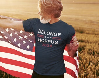 Puede incluir: Un hombre lleva una camiseta azul marino con el texto "DELONGE HOPPUS 2024" impreso en ella. Está sosteniendo una bandera estadounidense.