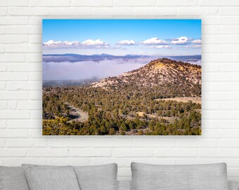 May include: An aerial view of a mountain range with a peak covered in trees and a layer of fog in the valley below. The sky is blue with white clouds.