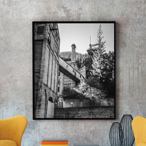 May include: Black and white photograph of an industrial building with a bridge connecting two sections. The photo is framed and hanging on a wall above two yellow armchairs and a coffee table.