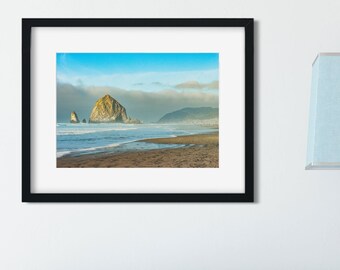 May include: A framed photograph of a rocky island off the coast of a beach. The island is a light brown color and is surrounded by blue water. The sky is a light blue color with some clouds.