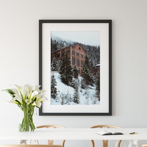 May include: A framed photograph of a brick building in a snowy forest. The building has multiple windows and a pitched roof. The photo is hanging on a white wall above a white table with four wooden chairs.