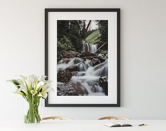 May include: A framed photograph of a waterfall cascading down a rocky hillside in a lush green forest. The photo is hanging on a white wall above a white table with four wooden chairs.