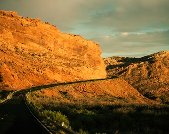 May include: A winding road through a desert landscape with red rock cliffs and a single car driving on the road.