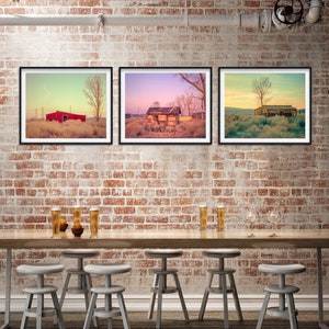Può includere: Tre fotografie incorniciate di edifici rustici con alberi e un cielo al tramonto appese a un muro di mattoni sopra un tavolo di legno con sei sgabelli da bar.
