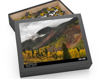 May include: A 500-piece jigsaw puzzle featuring a mountain landscape with a misty forest in autumn colors. The puzzle box is open, revealing the puzzle pieces inside. The box has the text "16x20 Puzzle 500 pcs Ages 9+"