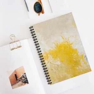 Op de afbeelding: Een spiraalgebonden notitieboek met een abstract ontwerp in wit en geel. Het ontwerp heeft een grote gele spatten met witte aderen.