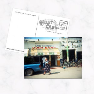 Könnte beinhalten: Eine Postkarte mit einem Foto eines blauen Lastwagens, der vor einem Gebäude mit einer rot-weißen Markise geparkt ist. Das Gebäude hat ein Schild mit der Aufschrift "Tacos Acorazados Dona Eva". Eine Frau geht vor dem Lastwagen entlang.