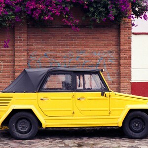 May include: A yellow vintage Volkswagen-style car with a black canvas roof, parked in front of a brick wall with graffiti. The car has a spare tire on the back and a black bumper.
