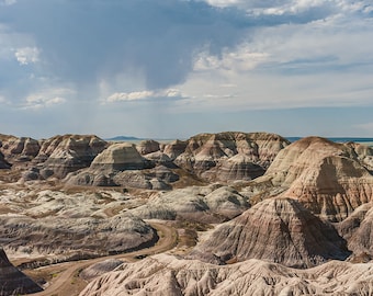May include: A panoramic view of a colourful, layered rock formation in a desert landscape. The rock formations are a variety of shades of brown, tan, white, and purple. The sky is blue with white clouds.