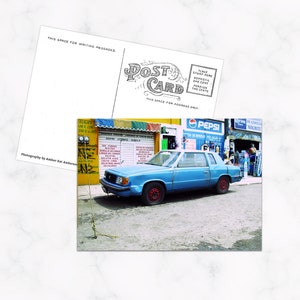 Könnte beinhalten: Eine Postkarte mit einem blauen Auto, das auf der Straße geparkt ist, vor einem Geschäft mit dem "PEPSI"-Logo. Die Rückseite der Postkarte ist sichtbar, mit Platz für eine Nachricht und Adresse. Die Vorderseite ist ein Foto.