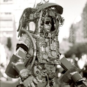 K&ouml;nnte beinhalten: Eine Person tr&auml;gt ein gro&szlig;es, kompliziertes Steampunk-Kost&uuml;m aus Metall, Zahnr&auml;dern und Rohren. Das Kost&uuml;m hat einen Helm mit Visier und verschiedenen Messger&auml;ten und Zifferbl&auml;ttern. Die Person steht vor einem verschwommenen Hintergrund.