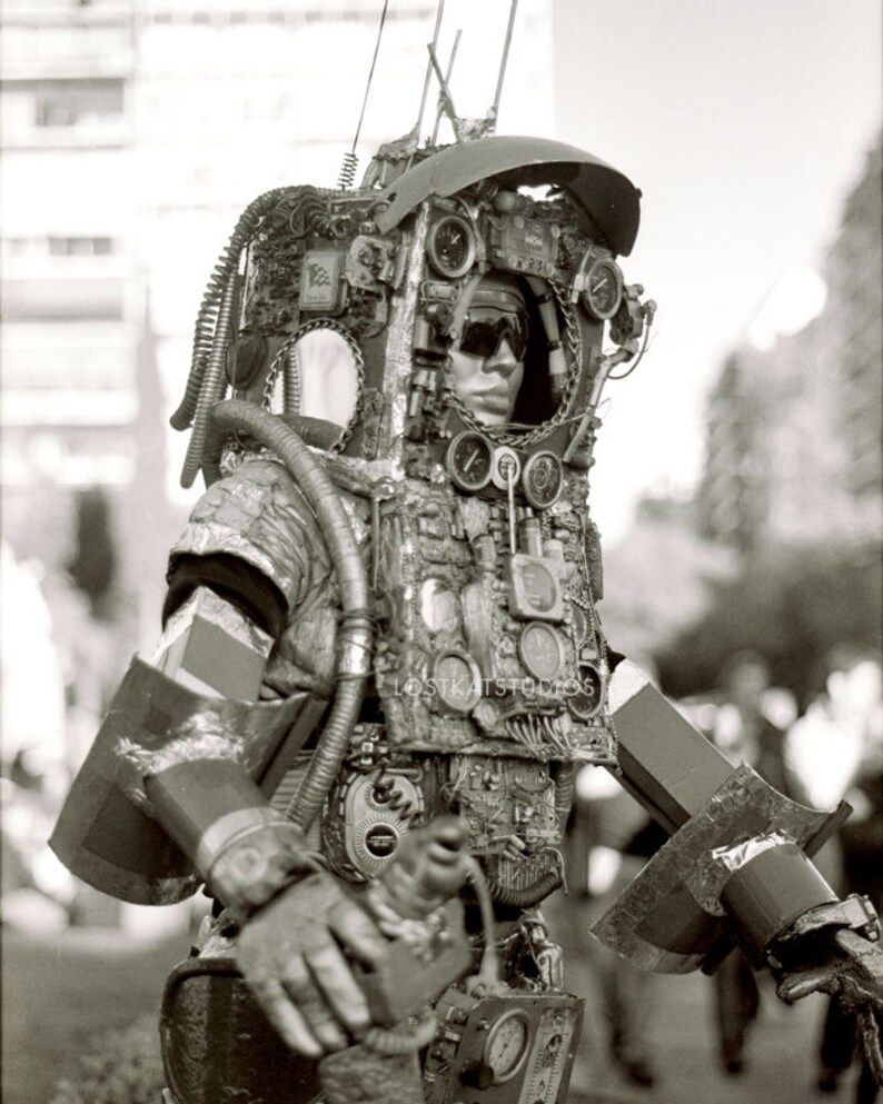 K&ouml;nnte beinhalten: Eine Person tr&auml;gt ein gro&szlig;es, kompliziertes Steampunk-Kost&uuml;m aus Metall, Zahnr&auml;dern und Rohren. Das Kost&uuml;m hat einen Helm mit Visier und verschiedenen Messger&auml;ten und Zifferbl&auml;ttern. Die Person steht vor einem verschwommenen Hintergrund.