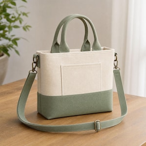 May include: A structured tote bag featuring a cream-colored upper section and a sage green lower base. The bag includes two top handles, a front exterior pocket, and a detachable, adjustable sage green shoulder strap with bronze-toned metal hardware.