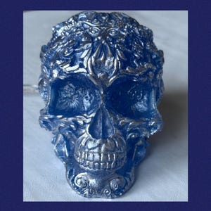 May include: A detailed, blue skull sculpture with silver accents. The skull features intricate floral and leaf patterns across the forehead and around the eye sockets. The teeth are clearly defined, and the overall design is gothic.