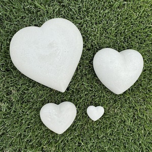 May include: Four white heart-shaped decorative stones of varying sizes are arranged on green grass. The largest heart is at the top left, with the smallest at the bottom right. The hearts have a textured surface.