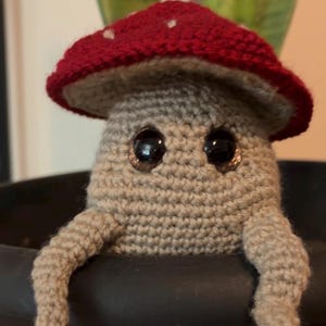 May include: A crocheted mushroom figure with a tan body and a red cap featuring small white spots. The figure has two large, dark, glossy eyes and two tan arms resting over the edge of a dark container. The texture is yarn-based and soft.