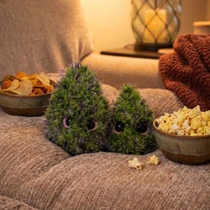 May include: Two green plush creatures with large eyes sit on a soft, textured surface, next to bowls of chips and popcorn. A cozy, relaxed scene with a red blanket in the background.