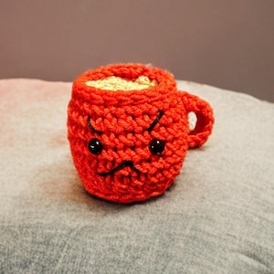 Könnte beinhalten: Eine gehäkelte rote Kaffeetasse mit einem mürrischen Gesicht und schwarzen Augen. Die Tasse ist mit einer beigefarbenen Substanz gefüllt.