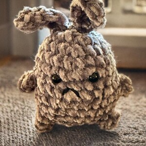 May include: A gray crocheted plush toy shaped like a bunny with a grumpy expression. The toy has black eyes and a small mouth.