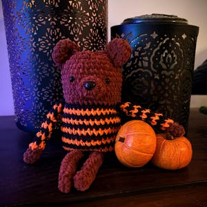 May include: A brown crocheted bear plush toy with a black and orange striped torso and arms. The bear has small round ears, a dark nose, and two small black eyes. Two small orange pumpkins are positioned next to the bear's lower body.