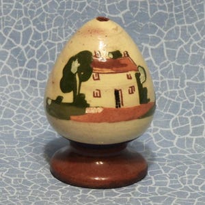 May include: A ceramic egg-shaped decorative item with a painted house scene. The egg is cream-colored with a brown base. The house has a red roof, and green trees are painted on either side.