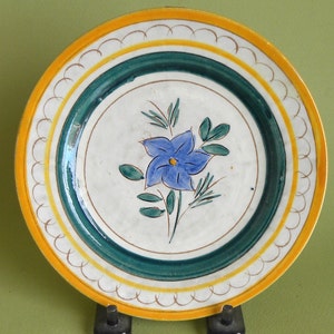 May include: A hand-painted ceramic plate with a floral design. The plate features a central blue flower with green leaves, surrounded by a dark green and yellow border. The rim has a scalloped edge. The plate is displayed on a black stand.
