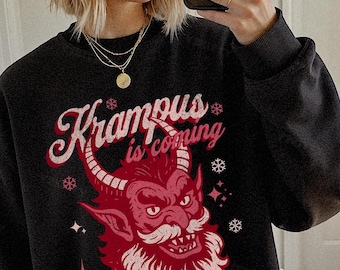 May include: Black sweatshirt with the text "Krampus is coming" in pink script and a red Krampus graphic. The Krampus graphic has a red face, horns, and beard. The person in the image is wearing a neon green beanie and sunglasses.