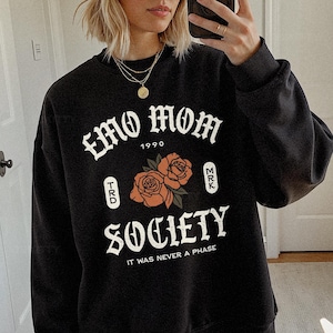 Puede incluir: Sudadera negra con el texto "EMO MOM 1990 SOCIETY" en fuente gótica blanca. El diseño incluye dos rosas naranjas y la frase "IT WAS NEVER A PHASE".