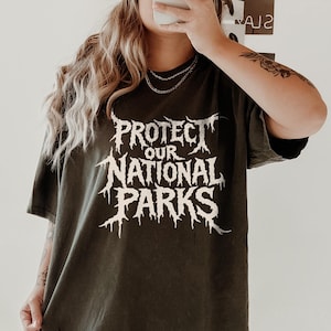 May include: An olive green t-shirt with the text "PROTECT OUR NATIONAL PARKS" in a white, stylized font. The shirt is oversized and worn with white sneakers and socks. The image is taken in front of a mirror.