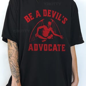 May include: Black t-shirt with the text "BE A DEVIL'S ADVOCATE" in red. The graphic features a red devil figure with wings. The shirt is a casual, oversized style.