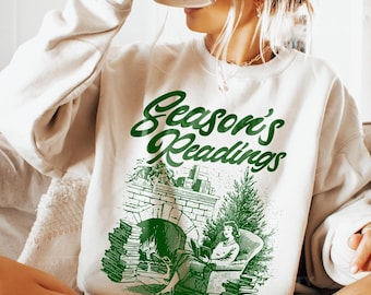 May include: Cream-colored sweatshirt with the text "Season's Readings" in green script. The graphic depicts a person reading by a fireplace. A black mug is held up to the face.