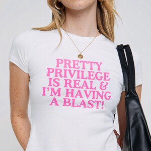 May include: White cropped t-shirt with pink text that reads "PRETTY PRIVILEGE IS REAL & I'M HAVING A BLAST!"