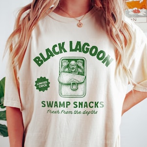 May include: Cream-colored t-shirt with a green graphic that reads "Black Lagoon" above an illustration of a creature in a can labeled "Swamp Snacks." Below, the text "Fresh from the depths" is printed in green.