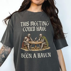 Può includere: T-shirt grigio scuro con la scritta "THIS MEETING COULD HAVE BEEN A RAVEN". La maglietta presenta un'illustrazione di due cavalieri seduti a un tavolo. Un cavaliere indossa un'armatura rossa e bianca, l'altro verde.