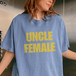 May include: A light blue t-shirt with the words "UNCLE FEMALE" printed in yellow. The shirt has short sleeves and a crew neck. The text is in a bold, sans-serif font.