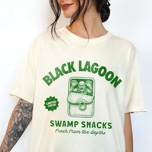 May include: Cream-colored t-shirt with a green graphic that reads "Black Lagoon" above an illustration of a creature in a can labeled "Swamp Snacks." The text "Fresh from the depths" is below the can. A "Limited Catch" badge is on the left.