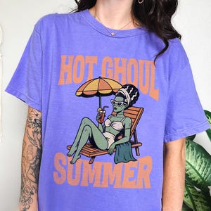 May include: A purple short-sleeve t-shirt featuring a graphic of a green-skinned figure in a white bikini lounging on a wooden chair under an orange umbrella, holding a drink. The text "HOT GHOUL SUMMER" is printed in orange above and below the graphic.
