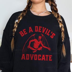 May include: Black sweatshirt with the text "BE A DEVIL'S ADVOCATE" in red. The design includes a red illustration of a devil figure. The sweatshirt is a casual, oversized style.