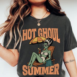 May include: A charcoal grey t-shirt featuring a graphic of a green-skinned character with a tall hairstyle, wearing sunglasses and a white bikini, lounging on a wooden chair under a yellow umbrella, with the text HOT GHOUL SUMMER in orange.