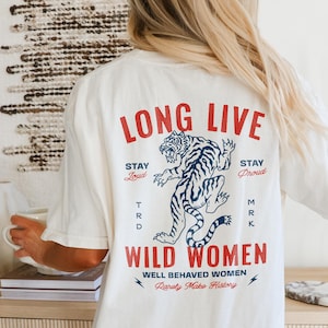 May include: Cream-colored t-shirt with the text "LONG LIVE WILD WOMEN" in red. A blue and white tiger graphic is centered below the text. The shirt also includes the phrases "Stay Loud", "Stay Proud", and "Well Behaved Women Rarely Make History."