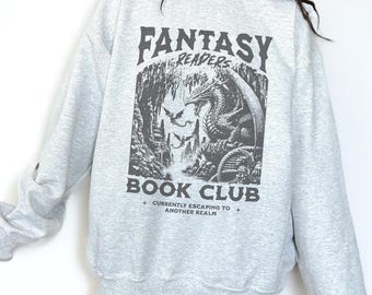 Puede incluir: Sudadera gris claro con la inscripción "FANTASY READERS BOOK CLUB" sobre una ilustración de un dragón y una cueva. El texto "CURRENTLY ESCAPING TO ANOTHER REALM" está debajo.